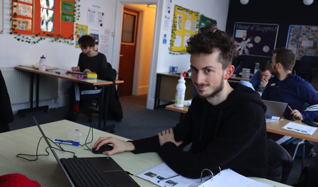 A young man sits in a classroom working on a computer. he smiles at the camera.