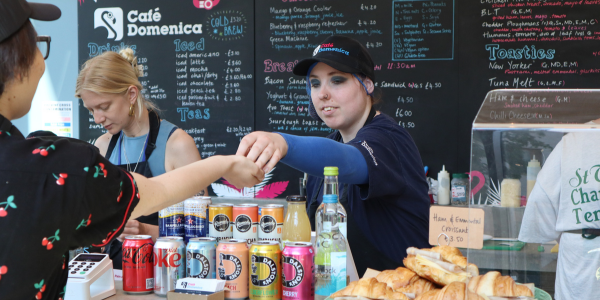 A woman serving a customer, wearing a black cap with Cafe Domenica logo on it and smiling