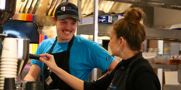 Two people stand together in a café. The young man is looking at their mentor and laughing. Both are wearing café Domenica uniforms.