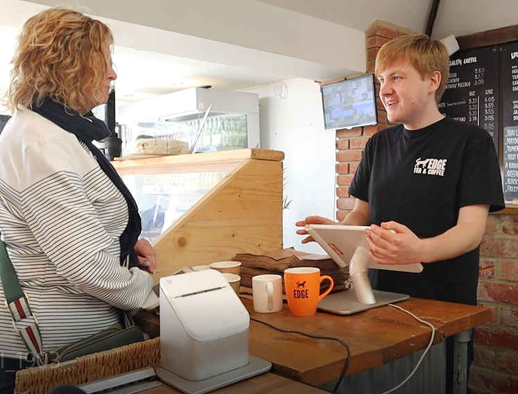 A young man is serving a woman at the till. He is smiling and wearing a black t-shirt with white EDGE Tea and Coffee logo.