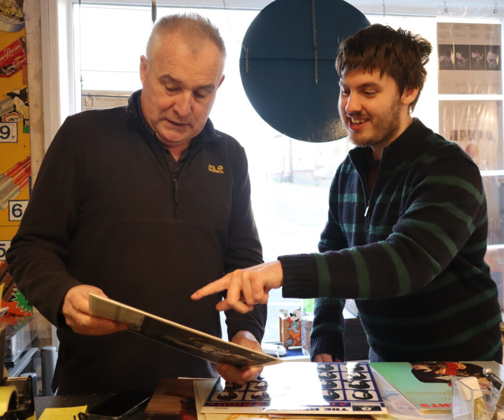 Two men talking, one is holding a vinyl record and the other one is pointing at it.