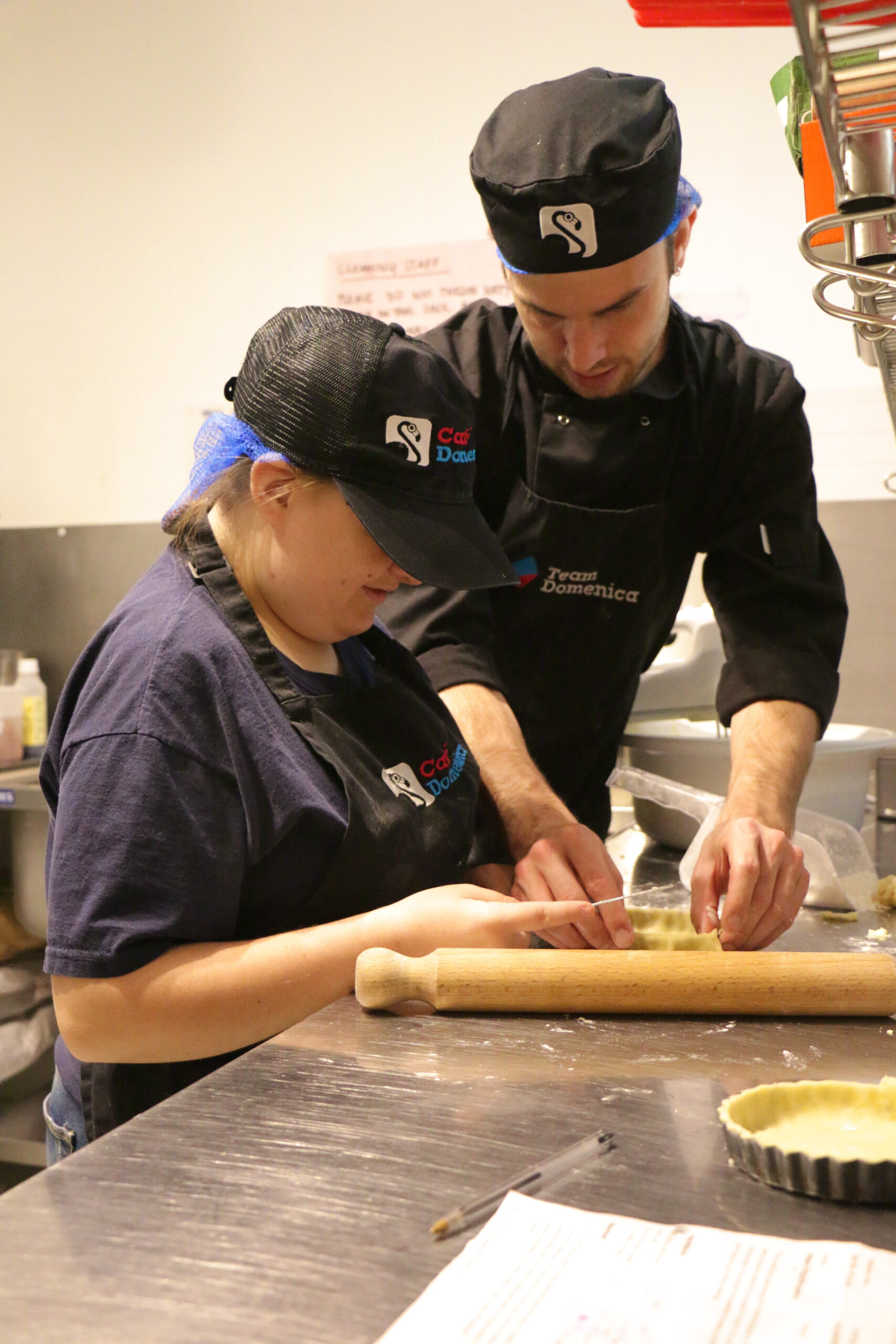 Two people work together to line and dish waith pastry. Both are wearing A Café Domenica hat and apron.