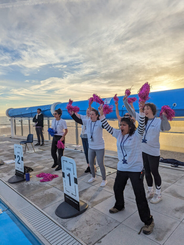 A group of people with pink shiny pom-poms in cheering poses with their arms up in the air. Background is a swimming pool and sunset shy at SEA LANES BRIGHTON.