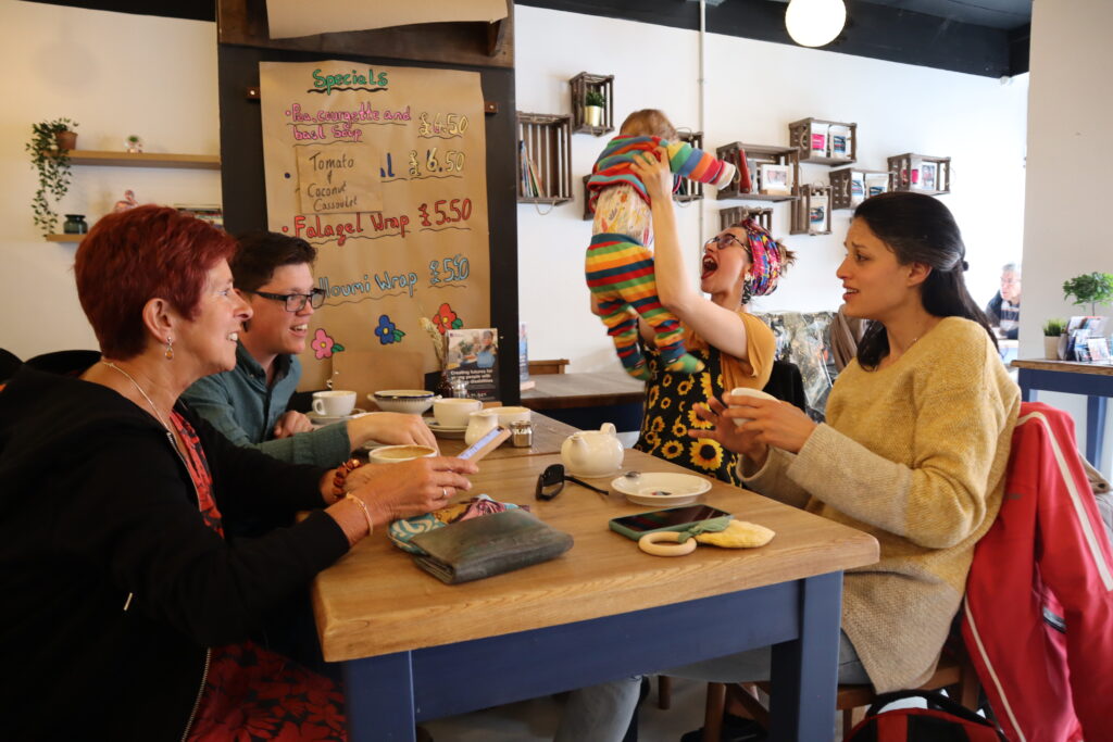 Image depicts three adults sat at a table, two are excitedly talking whilst the other makes their baby giggle by playing with them in the air.