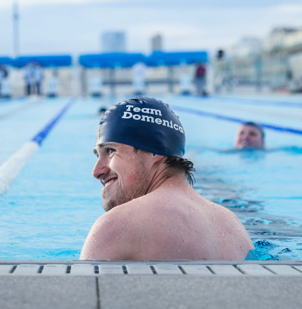 a swimmer smiles in the pool as he participates in Great Lengths. on his swim hat are the words Team Domenica.