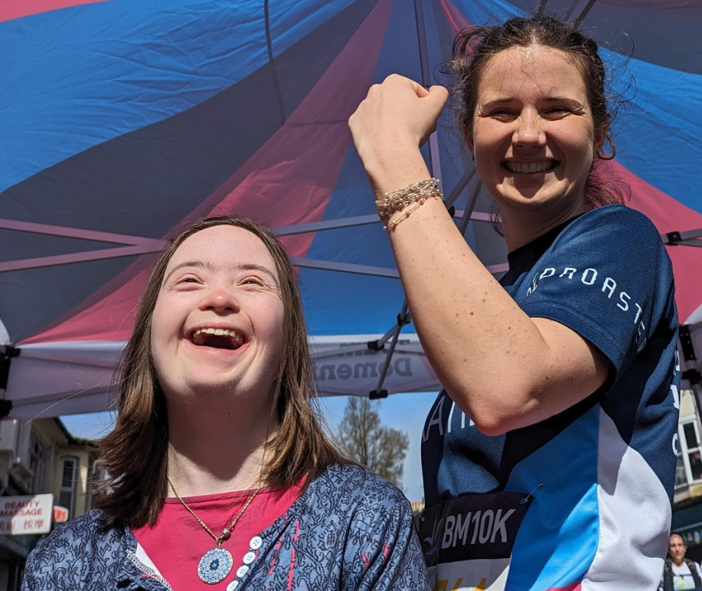 Two women stand next to one another. The person on the left (Sophie) is laughing and the person on the right (Katie) has a big grin as she flexes her arm muscles after completing the BM10k at Brighton Marathon Weekend 2024.