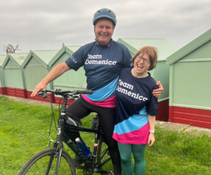 An image of Graham stood on a bike with his arm around his daughter, Betsy. They are both laughing and are wearing Team Domenica t-shirts.