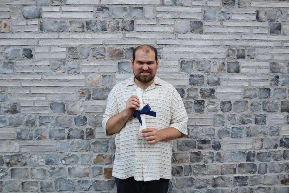 Man holding a graduation certificate standing in front of a wall and smiline